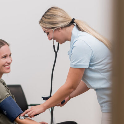 Blick durch die Tür, eine medizinische Fachangestellte misst den Blutdruck einer weiblichen Patientin