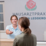 Medizinische Fachangestellte über gibt an der Rezeption einer Patientin eine Visitenkarte. Im Hintergrund sieht man das Praxislogo an der Wand und einen Blumenstrauß