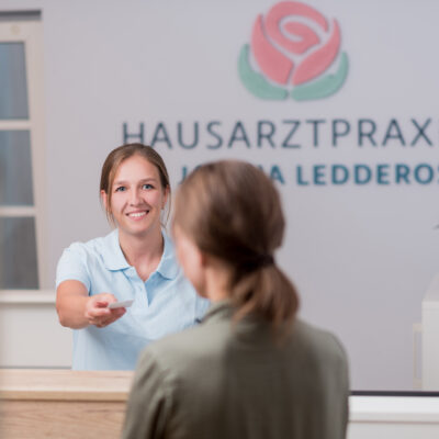 Medizinische Fachangestellte über gibt an der Rezeption einer Patientin eine Visitenkarte. Im Hintergrund sieht man das Praxislogo an der Wand und einen Blumenstrauß